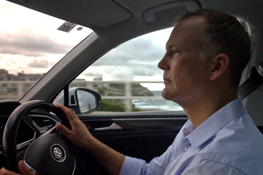 A man wearing a business shirt is sitting in the driver's seat of a car, his hands on the wheel. Out the window is a beach.