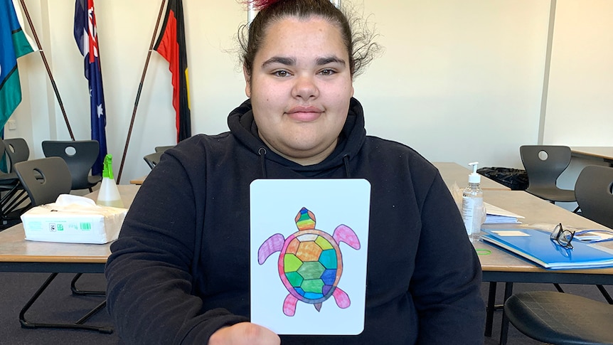Female High School students sits at desk, looking straight to camera, and holding an aboriginal designed card to the camera