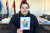 Female High School students sits at desk, looking straight to camera, and holding an aboriginal designed card to the camera