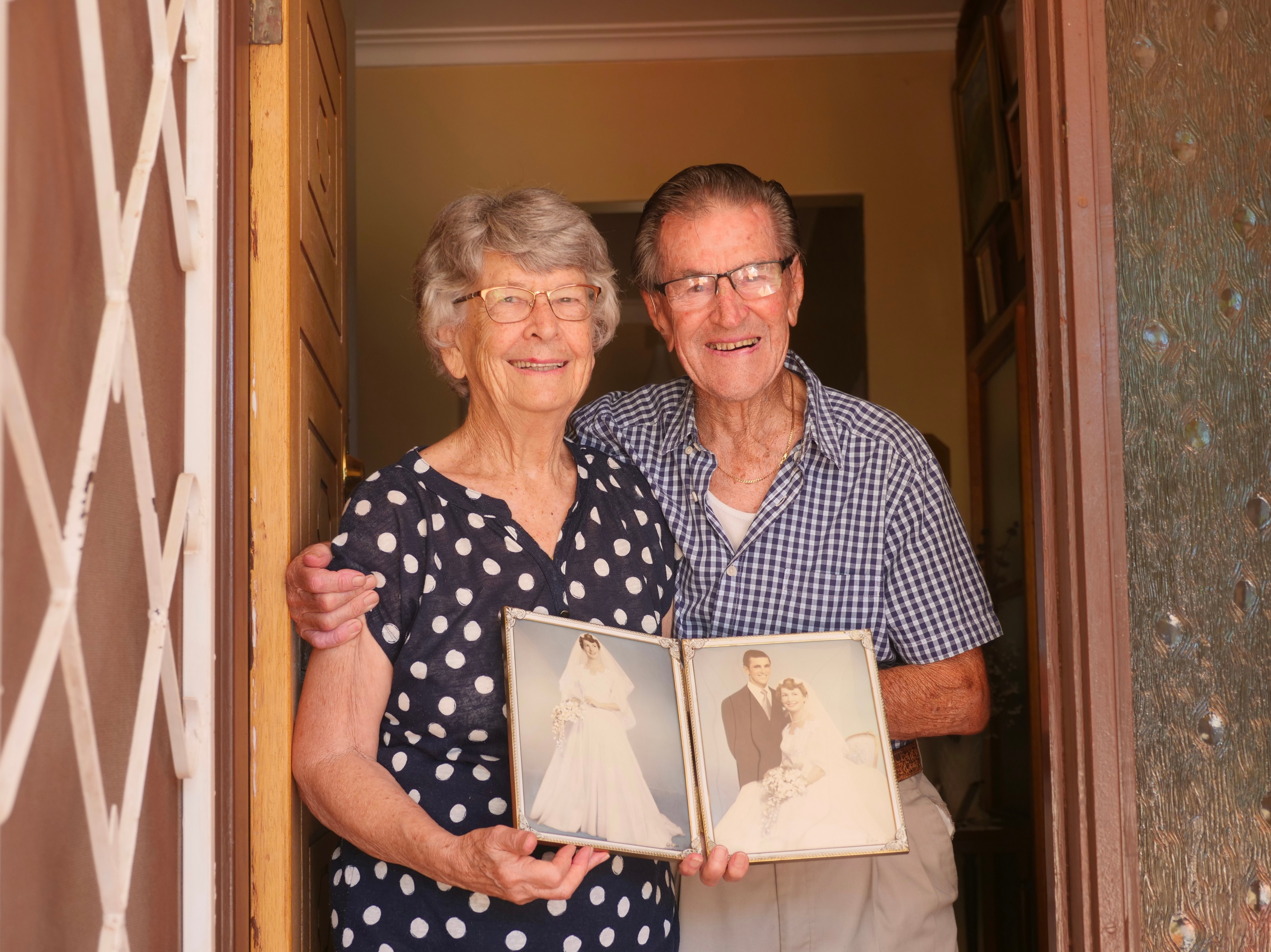 Una pareja mayor está parada en la puerta sosteniendo fotografías de boda.