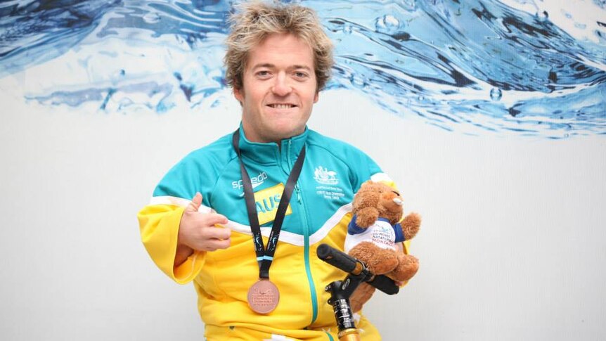 Man wearing Australian national swimming team tracksuit holds a stuffed beaver while posing with a medal around his neck.
