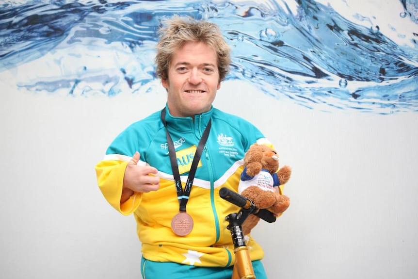 Man wearing Australian national swimming team tracksuit holds a stuffed beaver while posing with a medal around his neck.