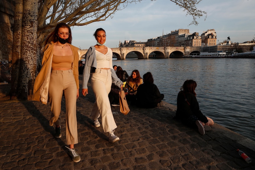 Two women walk with face masks on their chins along the Seine river in Paris