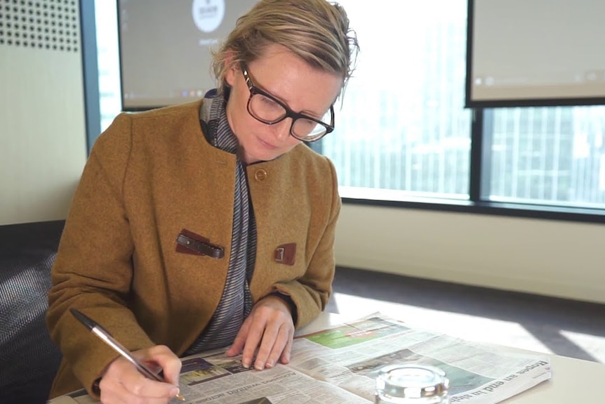 A woman sits at a table, marking a newspaper with notes.