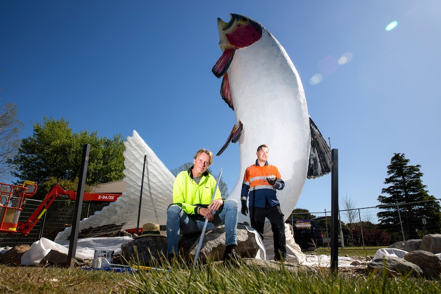 Two middle-aged men sitting in front of a big trout statue painted white and under restoration 