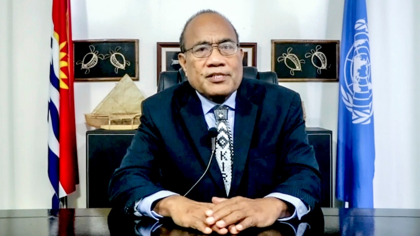 Taneti Maamau, wearing a suit, sits at a desk. Behind him are flags and paintings.