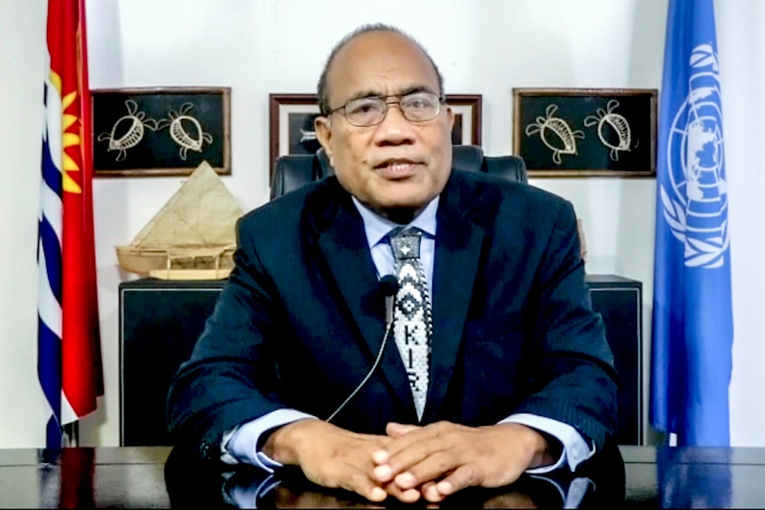 Taneti Maamau, wearing a suit, sits at a desk. Behind him are flags and paintings.