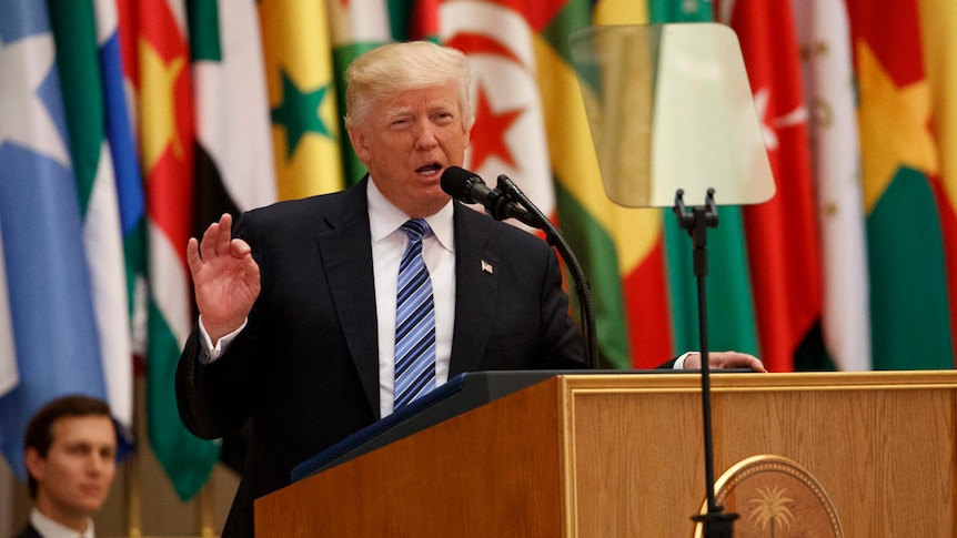 Donald Trump gestures as he speaks at a lectern. Flags are in the background.