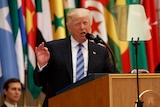 Donald Trump gestures as he speaks at a lectern. Flags are in the background.