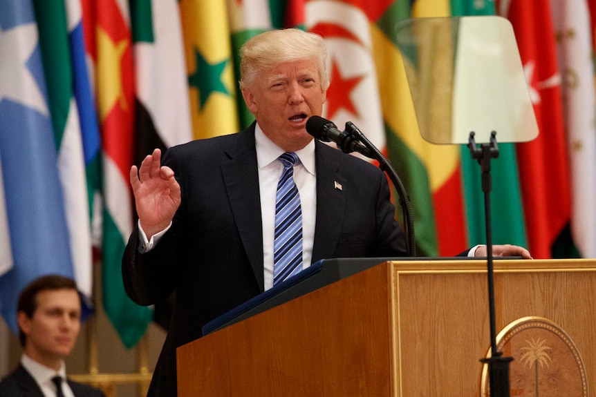 Donald Trump gestures as he speaks at a lectern. Flags are in the background.