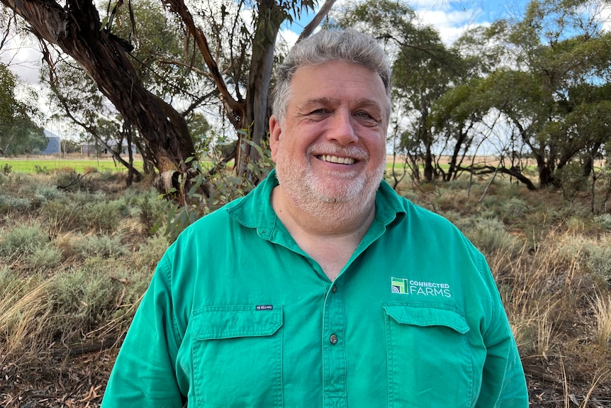 A smiling man with grey hair and a shirt beard, wearing a long-sleeve green work shirt, with Mallee trees in the backgorund.