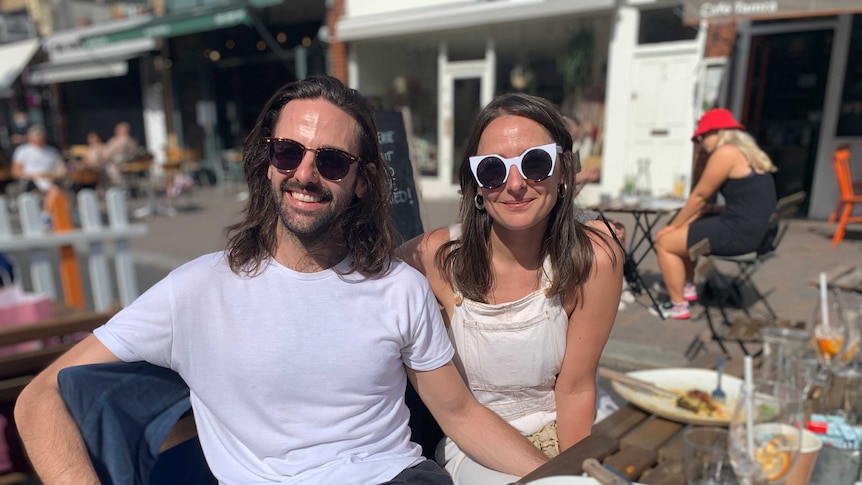 A man with long brown hair and wearing a white shirt with sunnies sits a girl wearing a dress and sunnies at a table outside.