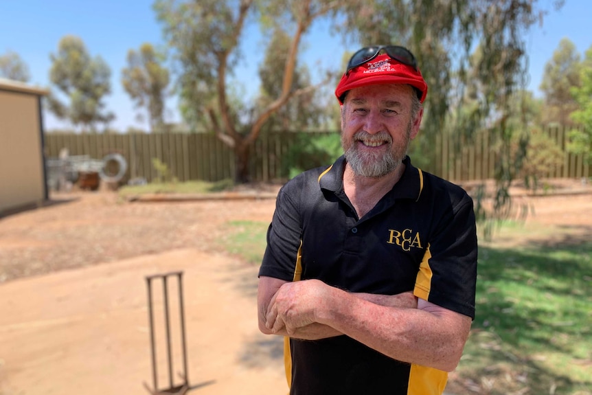Peter Kelly stands on a backyard cricket pitch with his arms crossed smiling with cricket stumps behind him