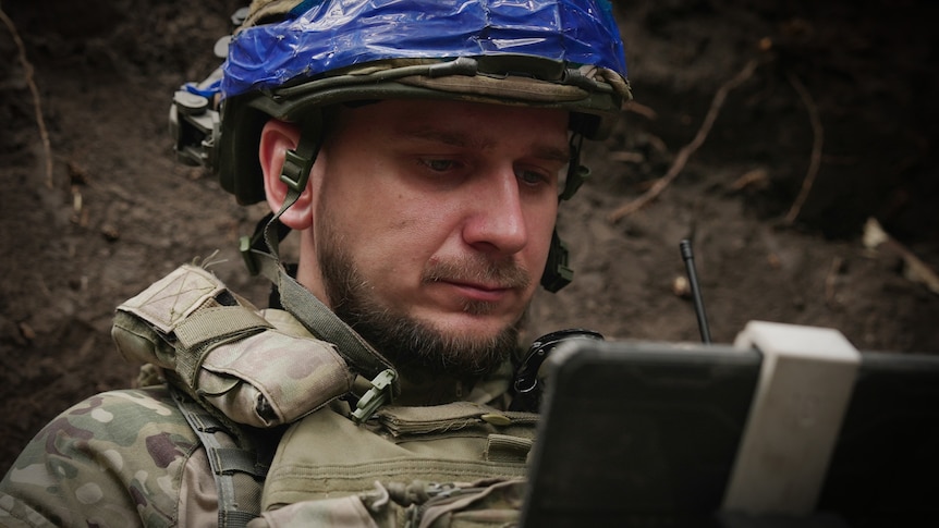 A man wearing a helmet and camouflage gear looks at his laptop.