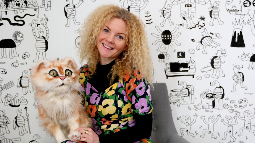 A blonde curly-haired blue-eyed woman holds a large ginger puppet cat, in story about puppetry helping dementia patients.
