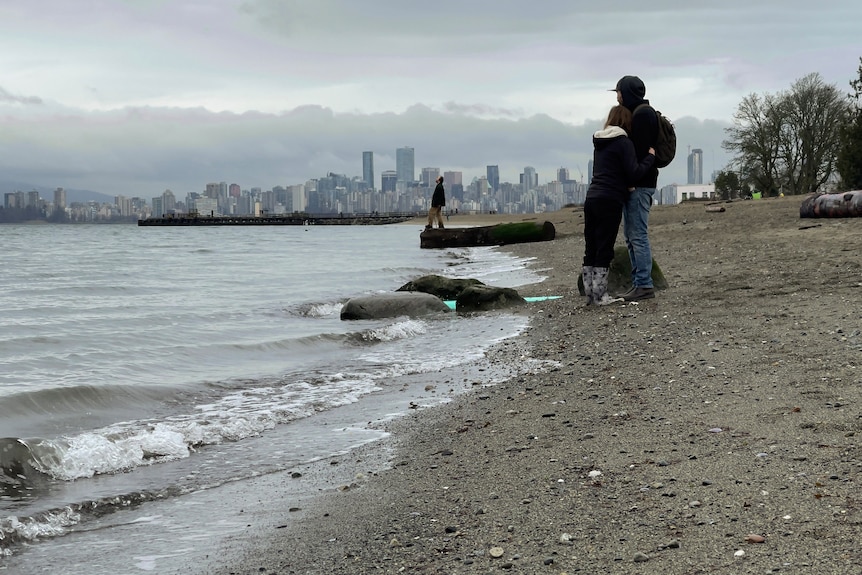 A couple hug at a beach in winter with Vancouver's skyline in the background