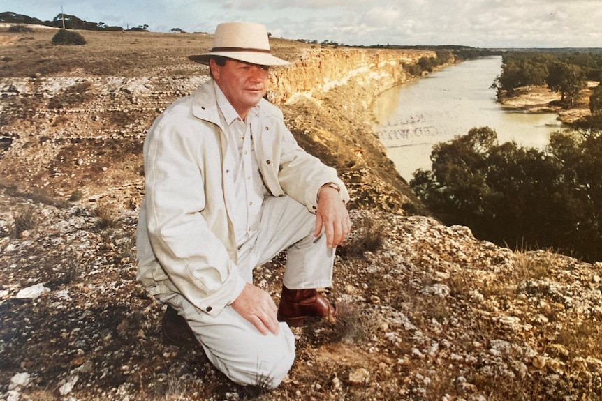  man in white pants, a white shirt and jacket and a white hat kneels in the countryside. 
