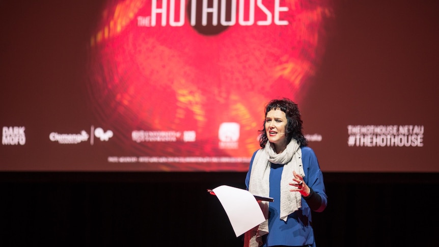 A woman with longish hair and a blue top talking on stage with a bright red backdrop with the words Hothouse on it