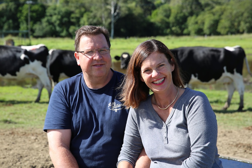 A couple smile at the camera with dairy cows behind hem.