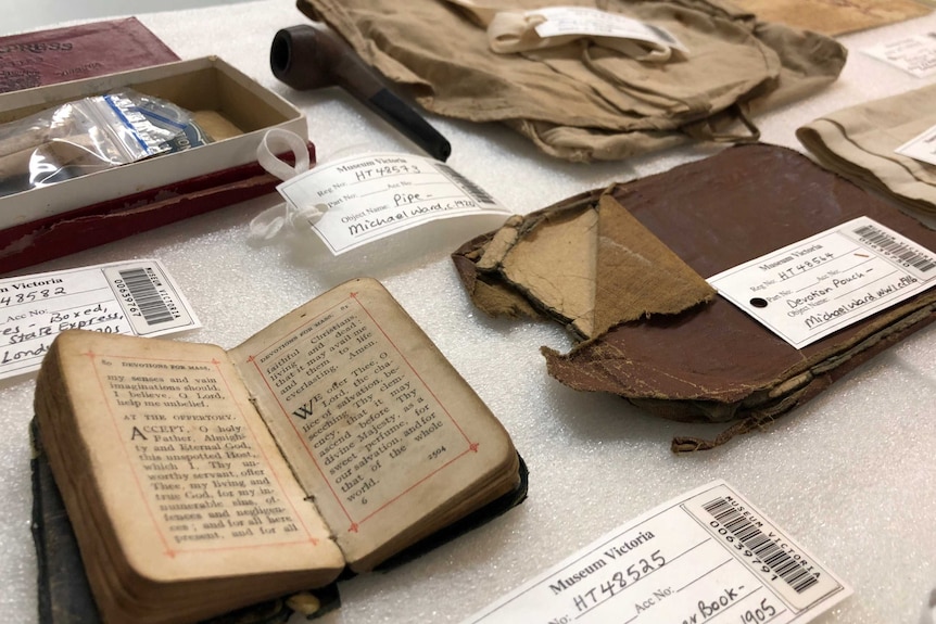 A prayer book, pipe, tattered devotion pouch and other old items are laid out on a table and labelled.