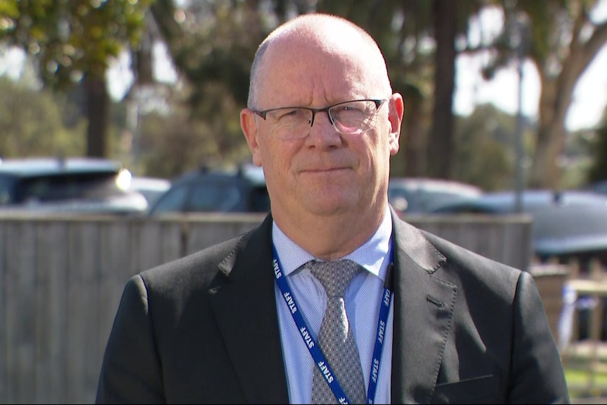 The detective has a bald head and wears steel rimmed glasses with a grey jacket and blue shirt and tie and stands outside.