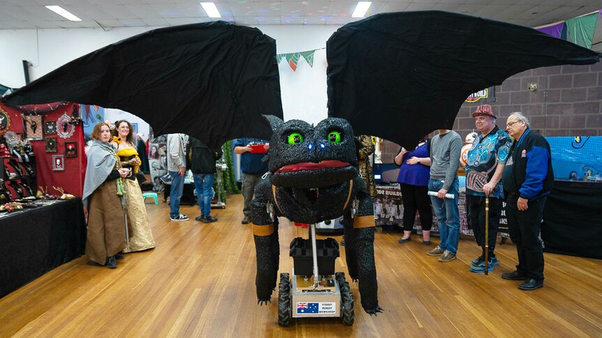 A black robot dragon with green eyes spreads his large wings while people look on.