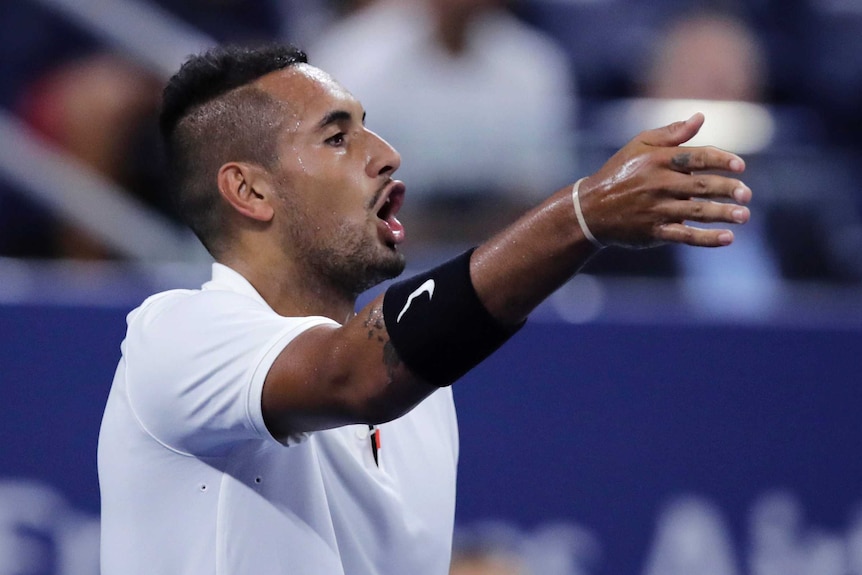 A male tennis player points his right arm out as he looks to his left as he speaks.