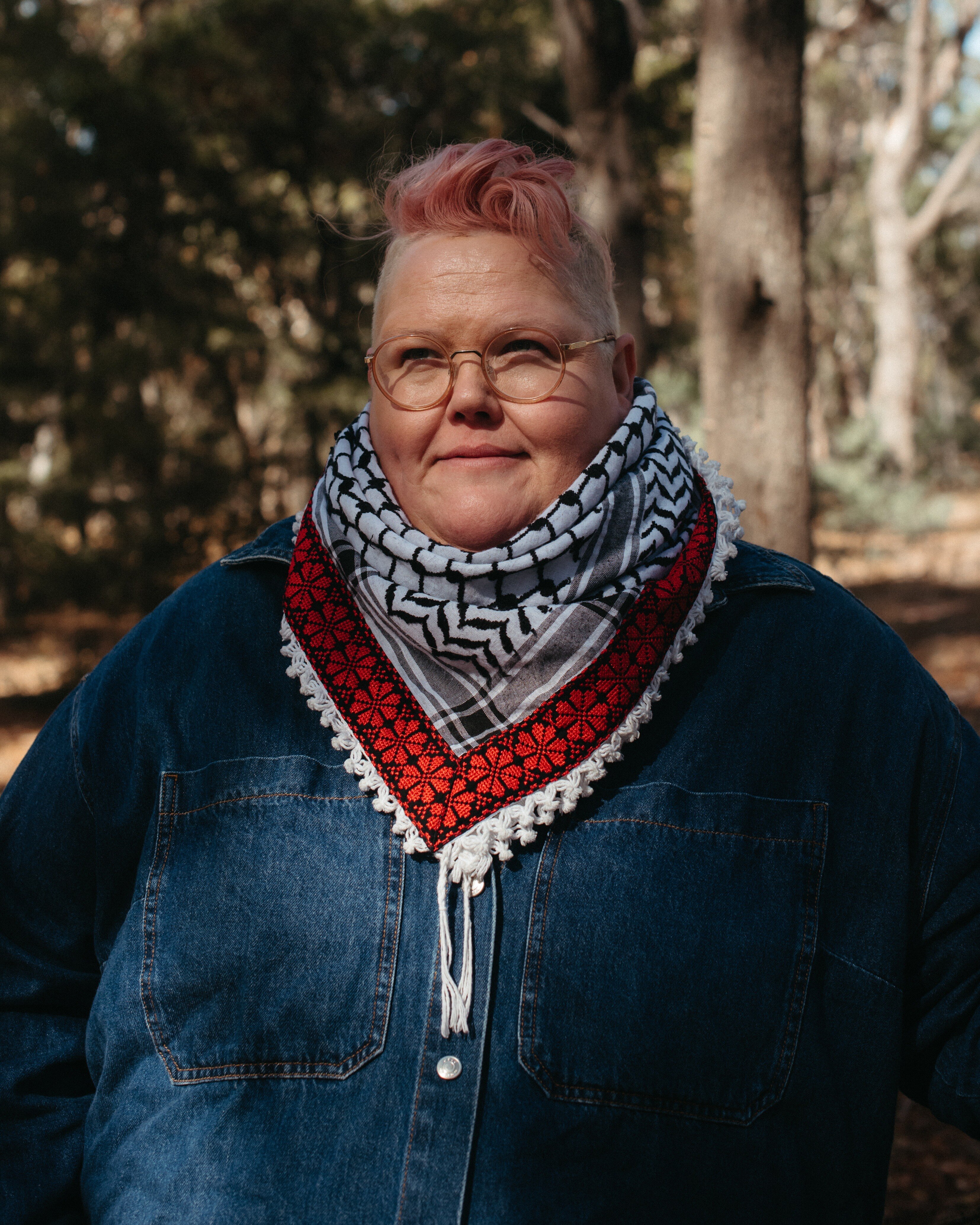 A close up shot of a person with pink hair and round glasses wearing a scarf and blue jumper