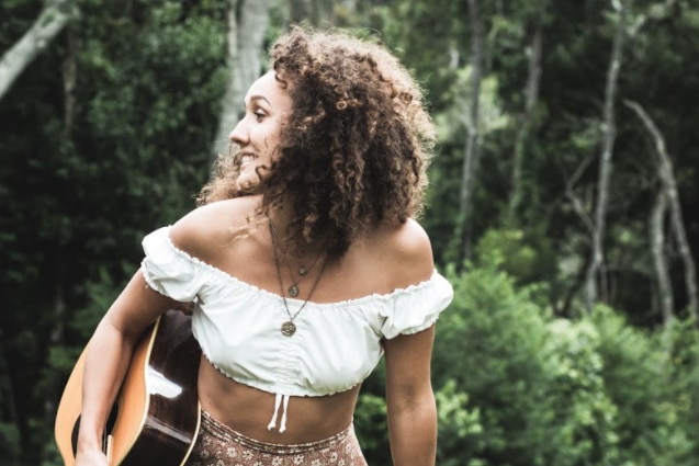 Musician NIDALA stands outside in a green leafy area with a guitar. She wears a white top and a necklace