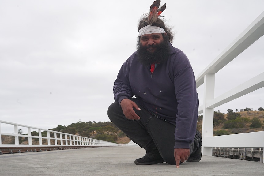An Indigenous man wearing a headband and feathers points to the deck of a causeway