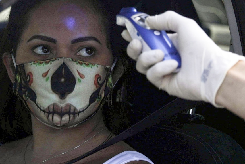 A woman driving a car with a face mask has her temperature taken by a health worker