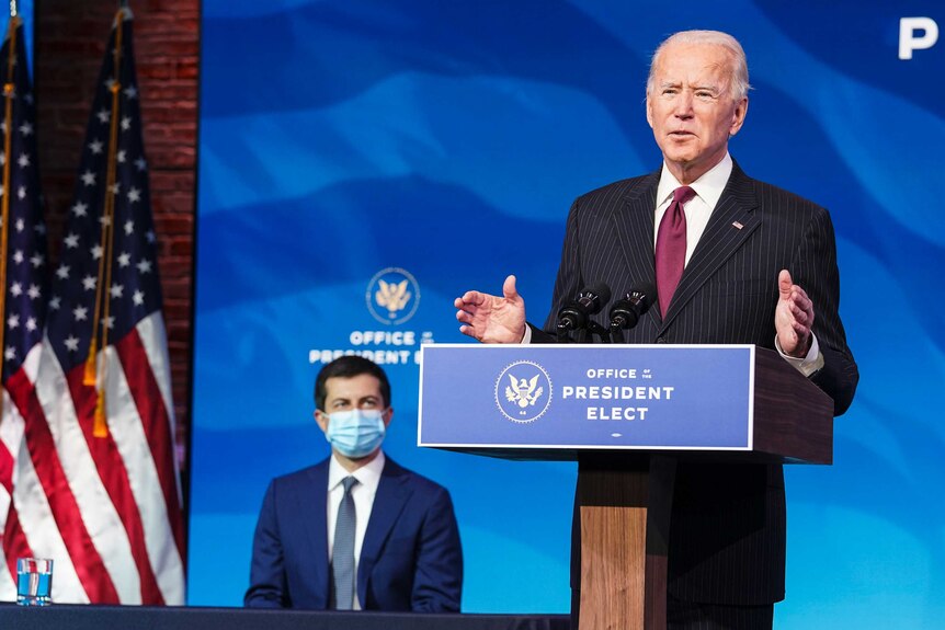 Pete Buttigieg is seated behind Joe Biden as he speaks on stage