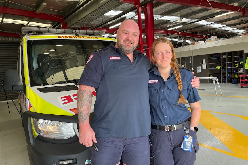 A very tall male paramedic stands next to a regular sized red headed female paramedic with a plait in front of an ambulance.