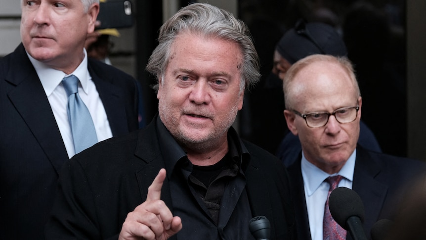 Steve Bannon is pictured holding up one finger while speaking into a microphone as two suited men stand behind him.