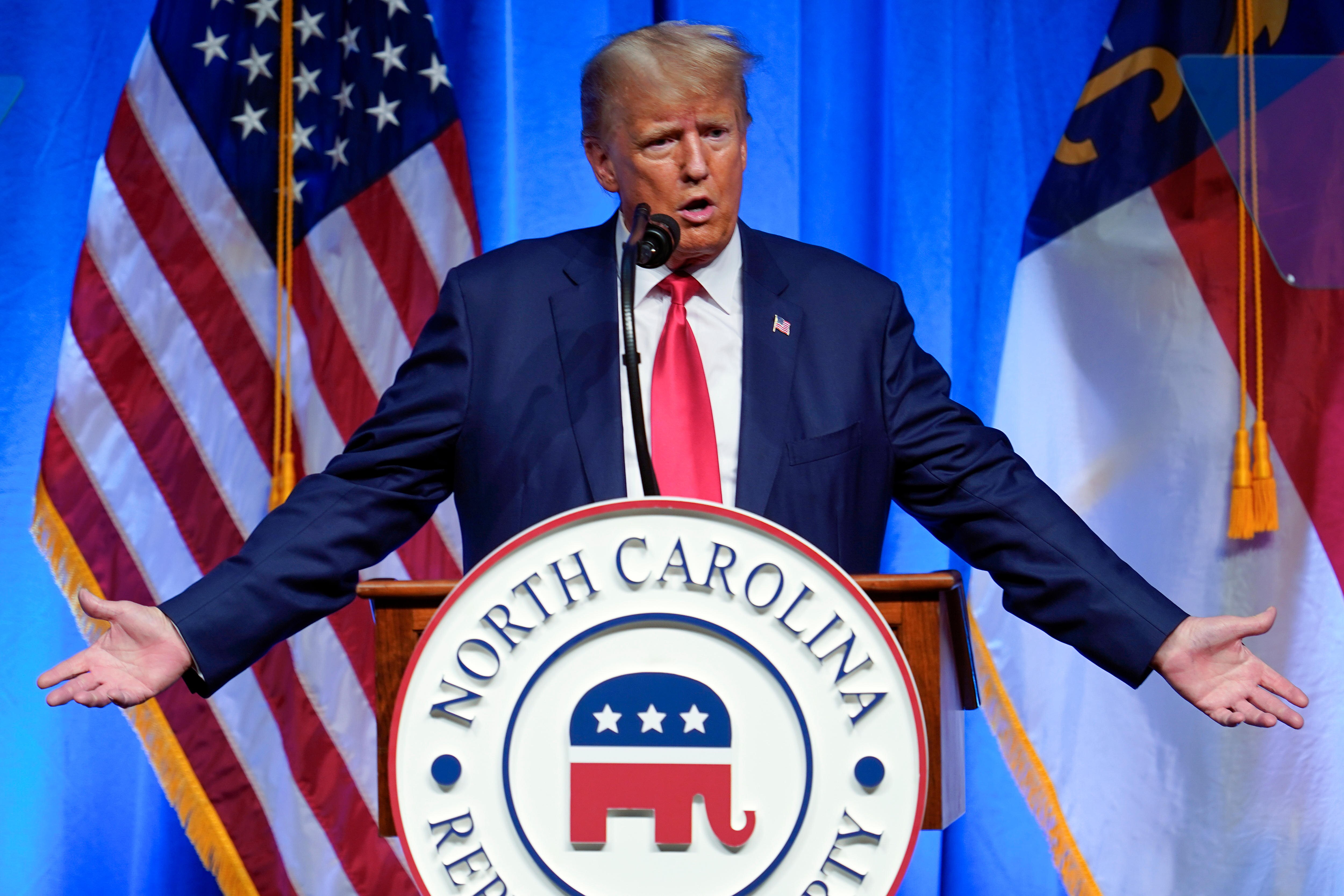 Donald Trump holds his arms outwards and low, with palms forward, in a questioning gesture as he gives a speech.