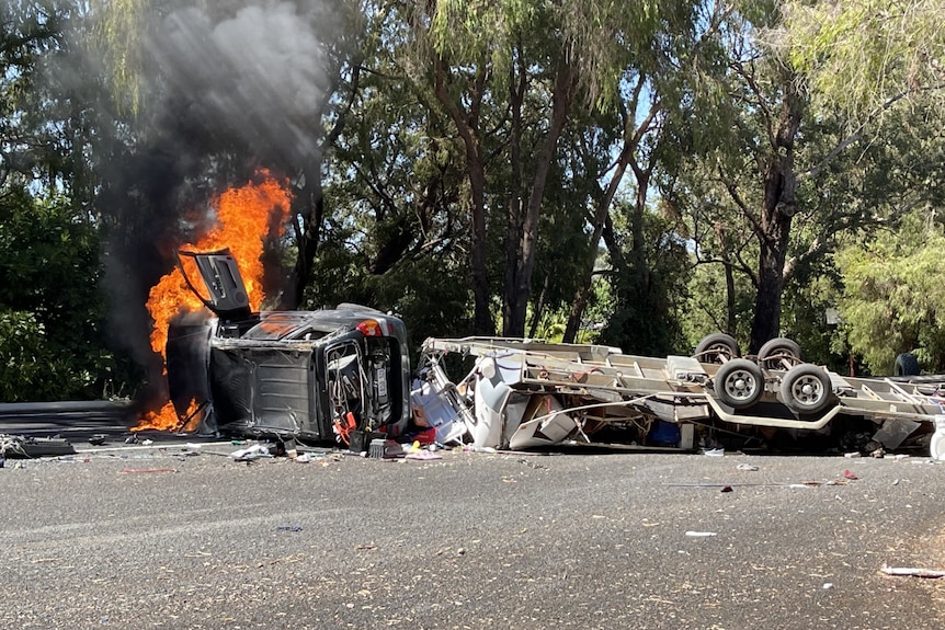 A car lays on it's side with smoke and flames flying into the air while a towed caravan lays wrecked on the road next to it.