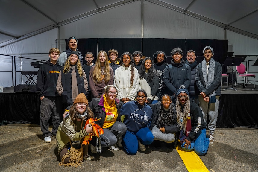 A group of teenagers pose for a photo in front of an elevated stage in a large marquee.