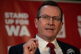 A close-up shot of Mark McGowan wearing glasses standing at a lectern in front of WA Labor party signage.