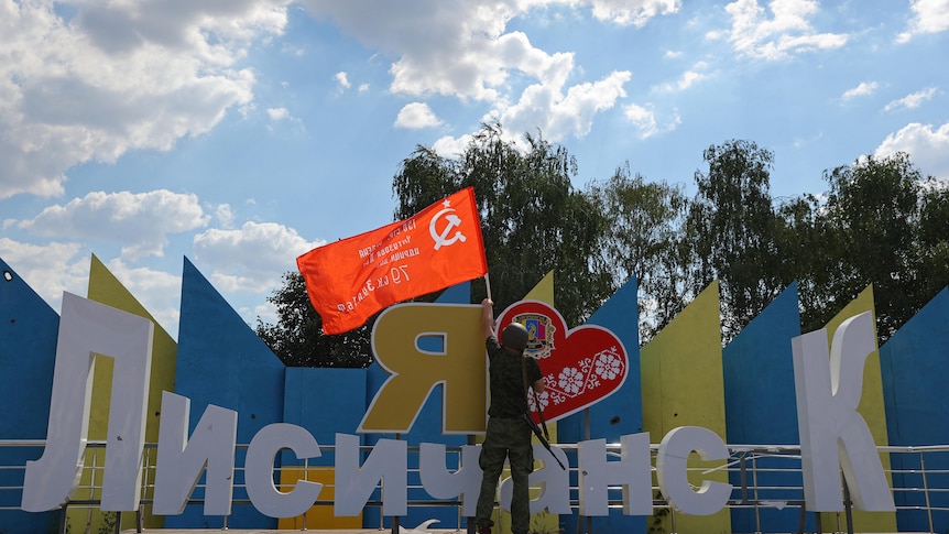A man raises a Soviet-era flag at a yellow and blue monument that says I love Lysychansk
