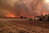 Fire is seen on the horizon with smoke covering the sky, water tanks and a home sit on dried land in the forefront.