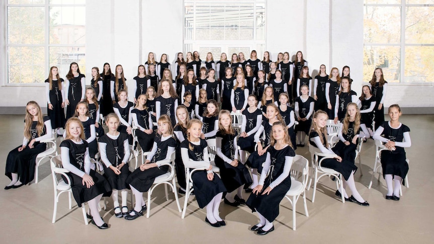 A girls' choir seated and standing in a white room with large windows.