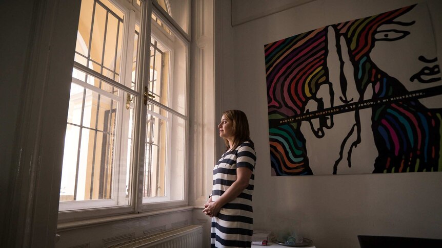 A woman in a black and white striped dress looks out of a large arched window in a house with high ceilings.