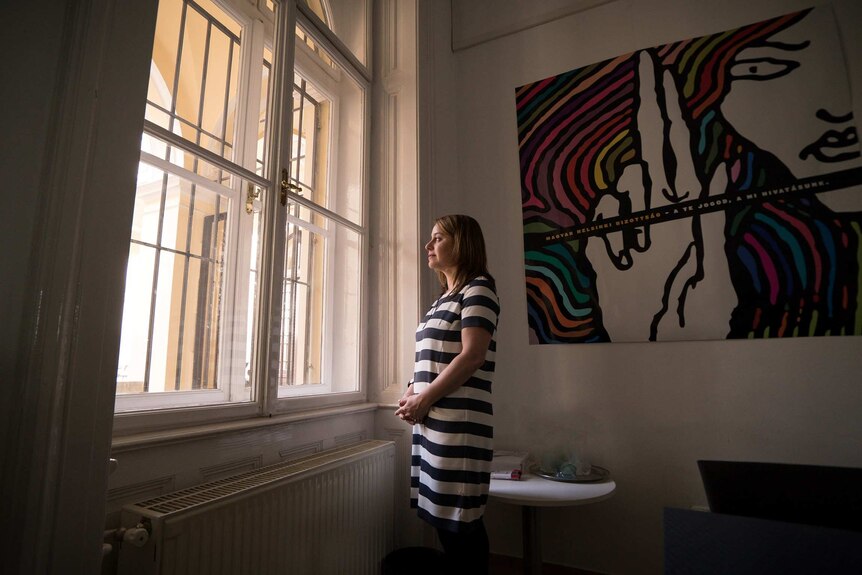 A woman in a black and white striped dress looks out of a large arched window in a house with high ceilings.