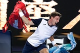 A man wearing tennis clothes grimaces as he holds a red bag above his head in one hand and a blue towel in the other
