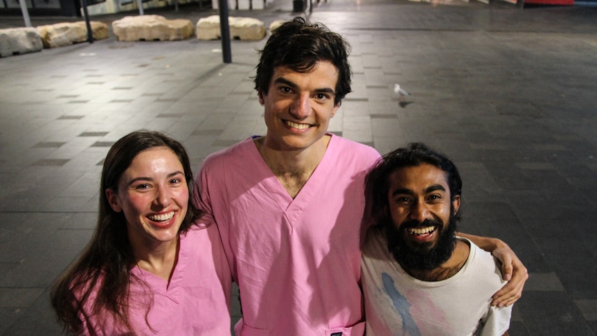 A doctor and a medical student wearing pink scrubs, with another medical student wearing a t-shirt, in a street at night