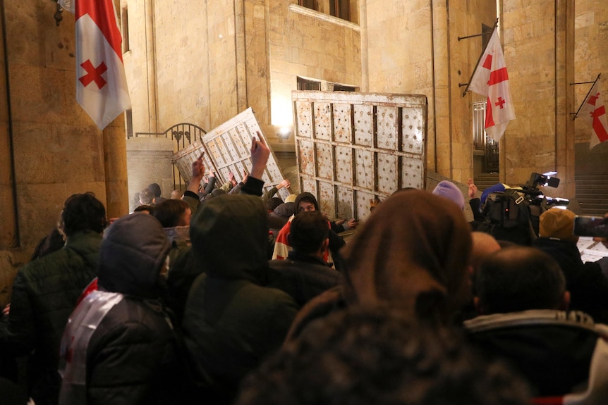 A group of people are in the process of tipping over a tall silver wall in front of a sandstone building