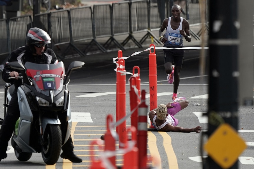 A marathon runner lies on the ground off the road, watching his nearest competitor running towards him during a big race. 