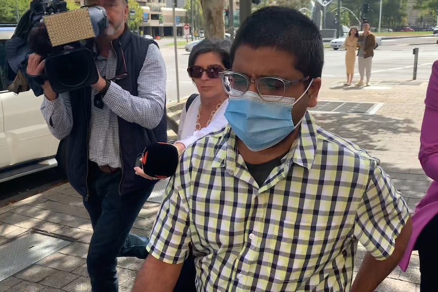 A man in a plaid yellow shirt, glasses and face mask walks out of court being questioned by reporters. 