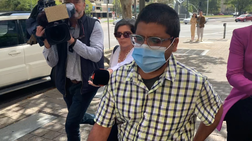 A man in a plaid yellow shirt, glasses and face mask walks out of court being questioned by reporters. 