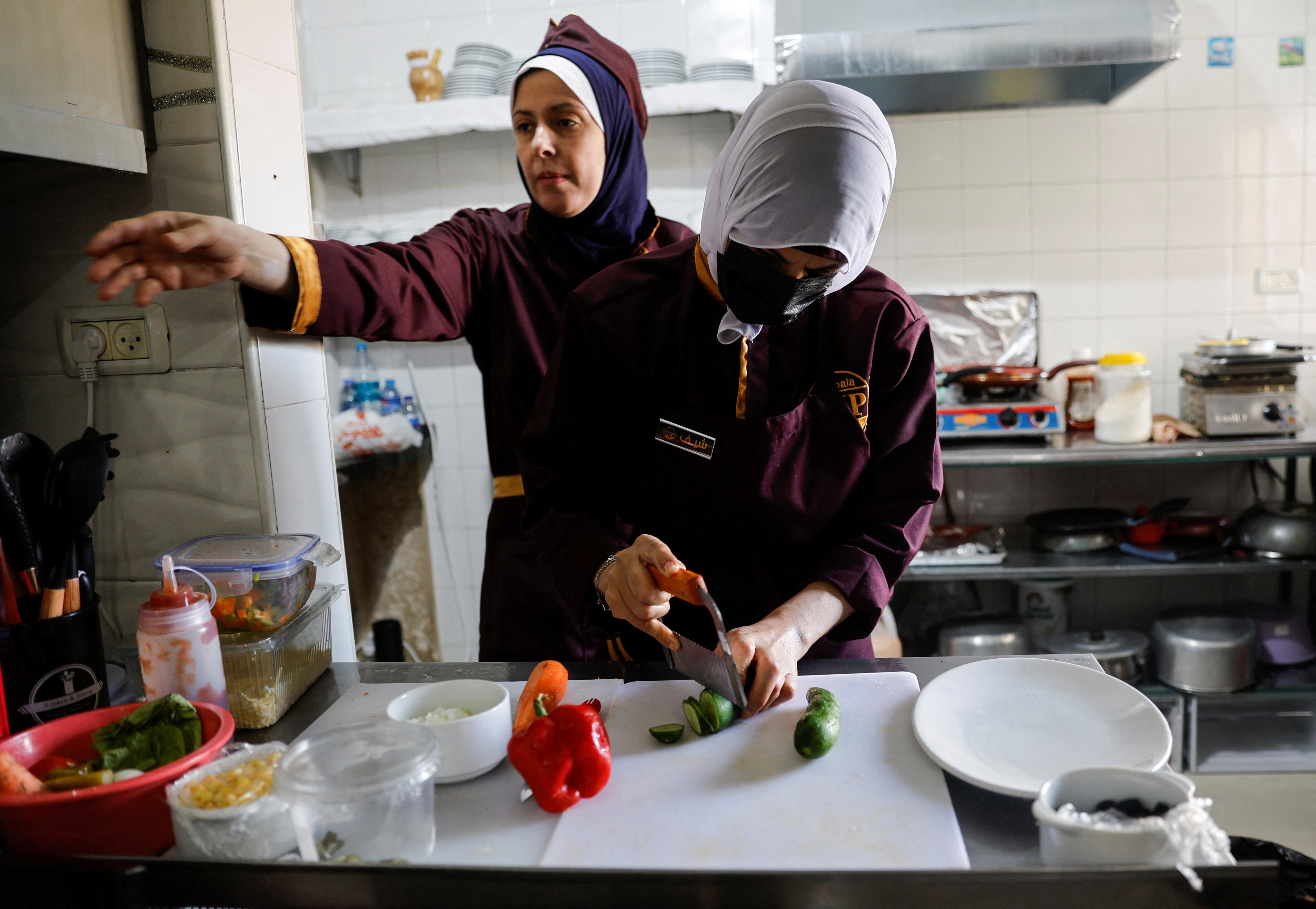 A woman reaches her arm out while another cuts up a cucumber